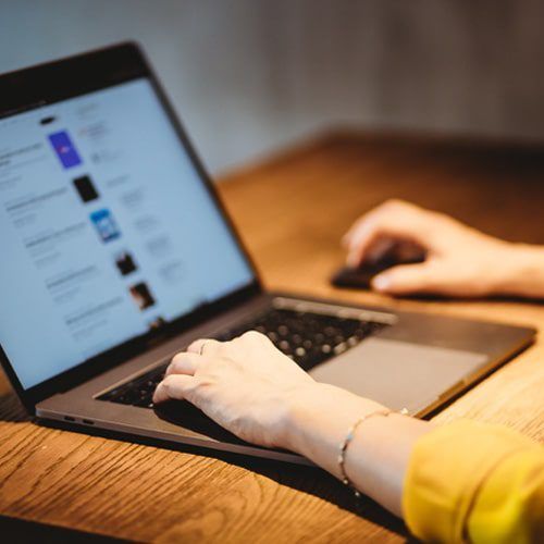 Person typing on a laptop with one hand and using a mouse with the other on a wooden desk.