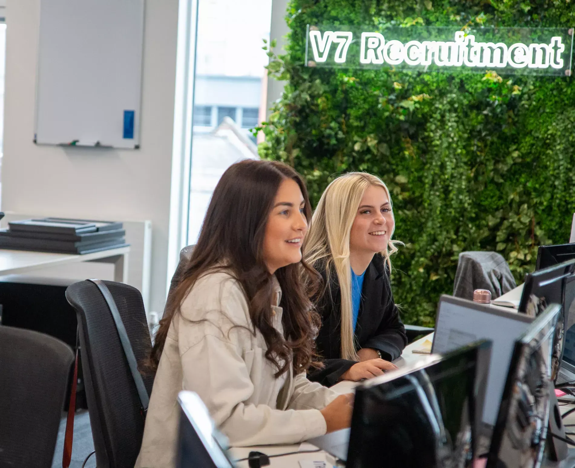 Two women smiling, working at computers in an office with a "V7 Recruitment" sign and greenery.