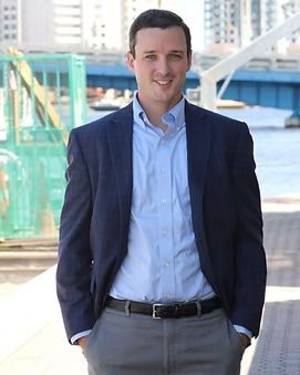 A man with dark hair and a blue blazer stands near a waterfront, smiling with hands in his pockets.