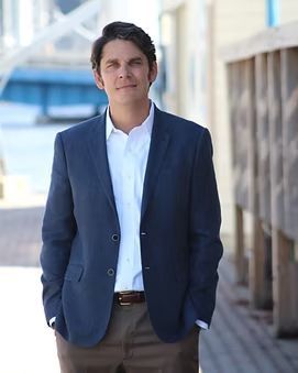 Man in blue blazer and white shirt, standing outdoors with hands in pockets.