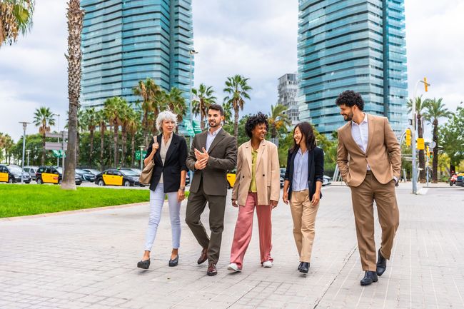 A diverse group of five people in business attire walking on a city path, modern buildings in the background.