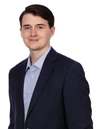 A young man in a suit and blue shirt is standing in front of a white background.