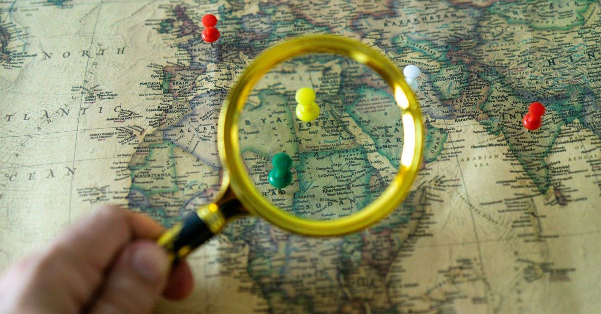 A person is holding a magnifying glass over a map with pins on it.