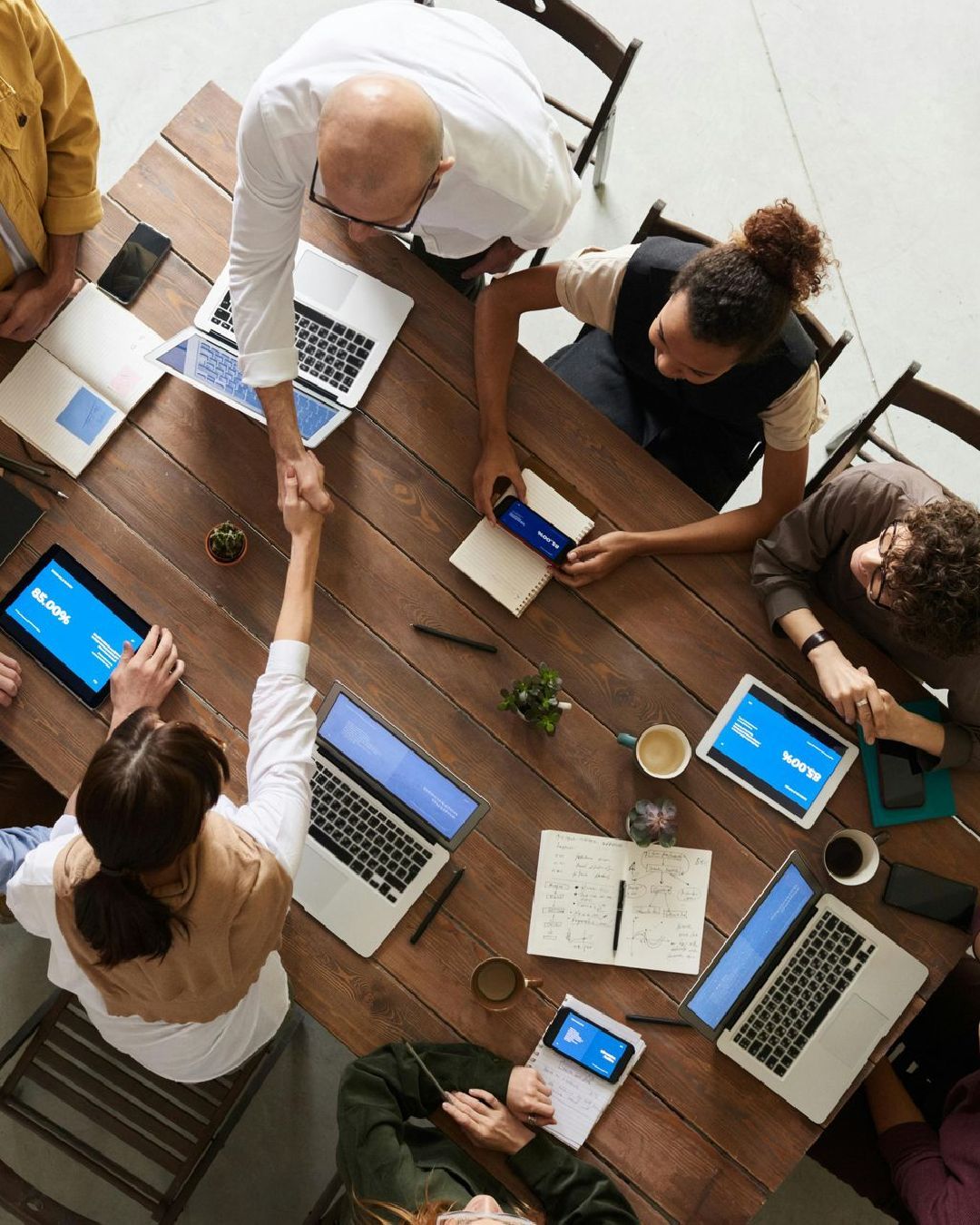 A group of people are sitting around a wooden table with laptops and tablets.