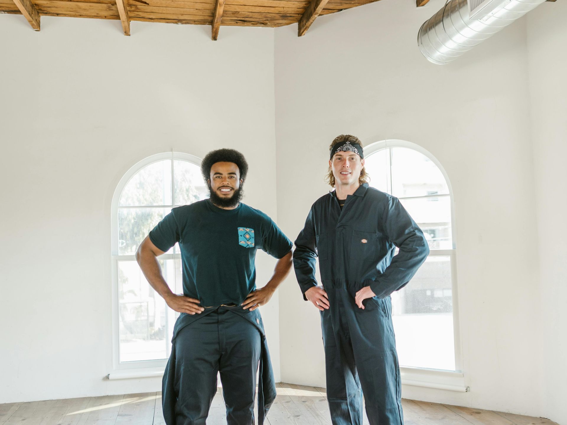 Two men are standing next to each other in an empty room with their hands on their hips.