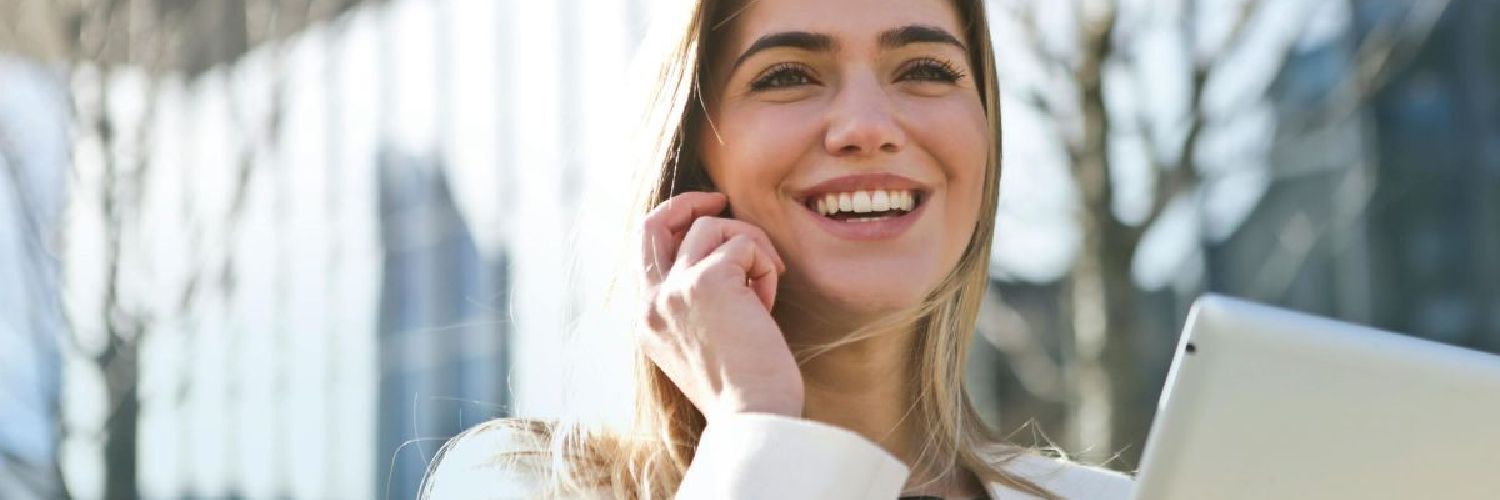 A woman is smiling while talking on a cell phone and holding a tablet.