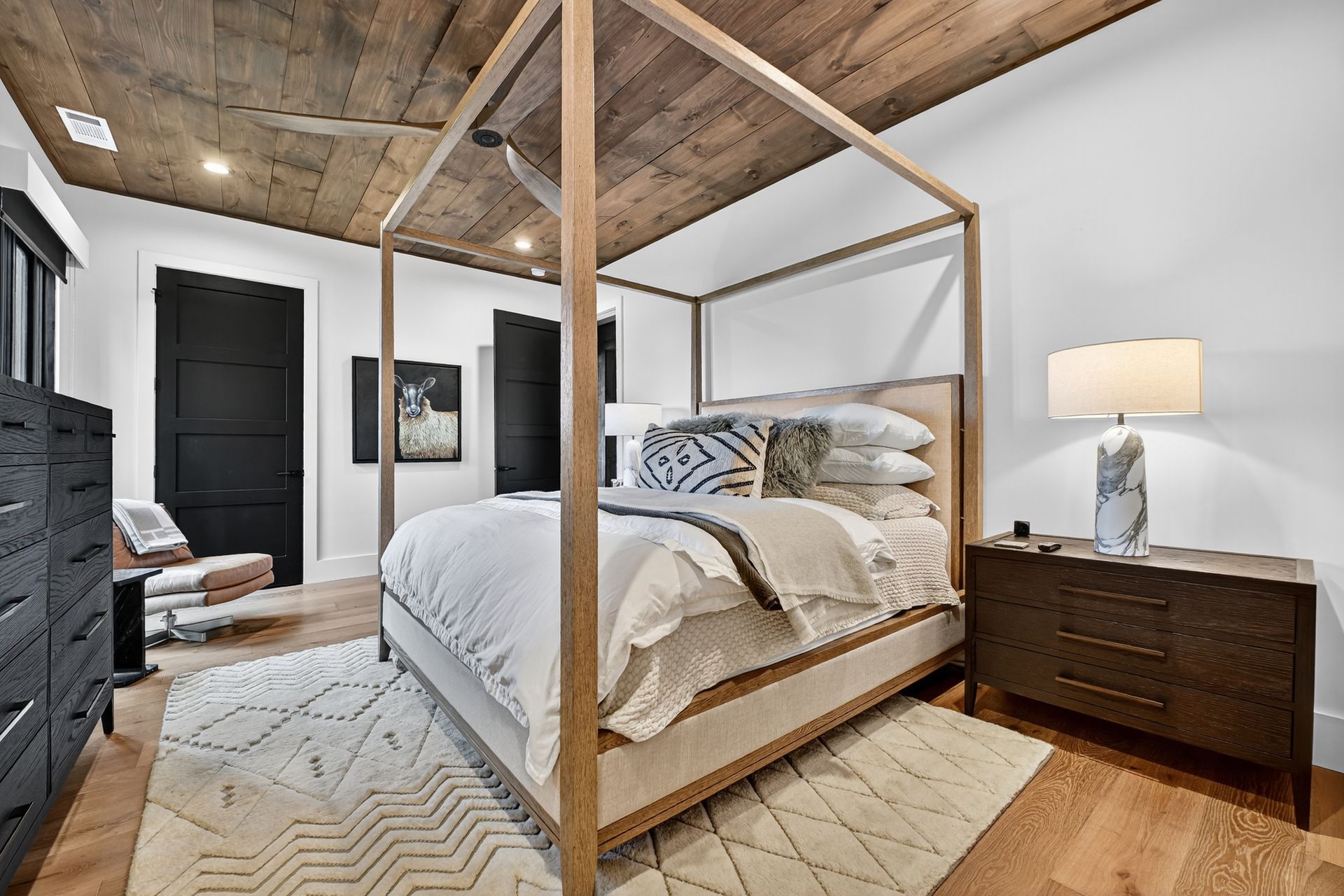 Bedroom with canopy bed, wood ceiling and floors, white bedding, rug, and black accent doors.