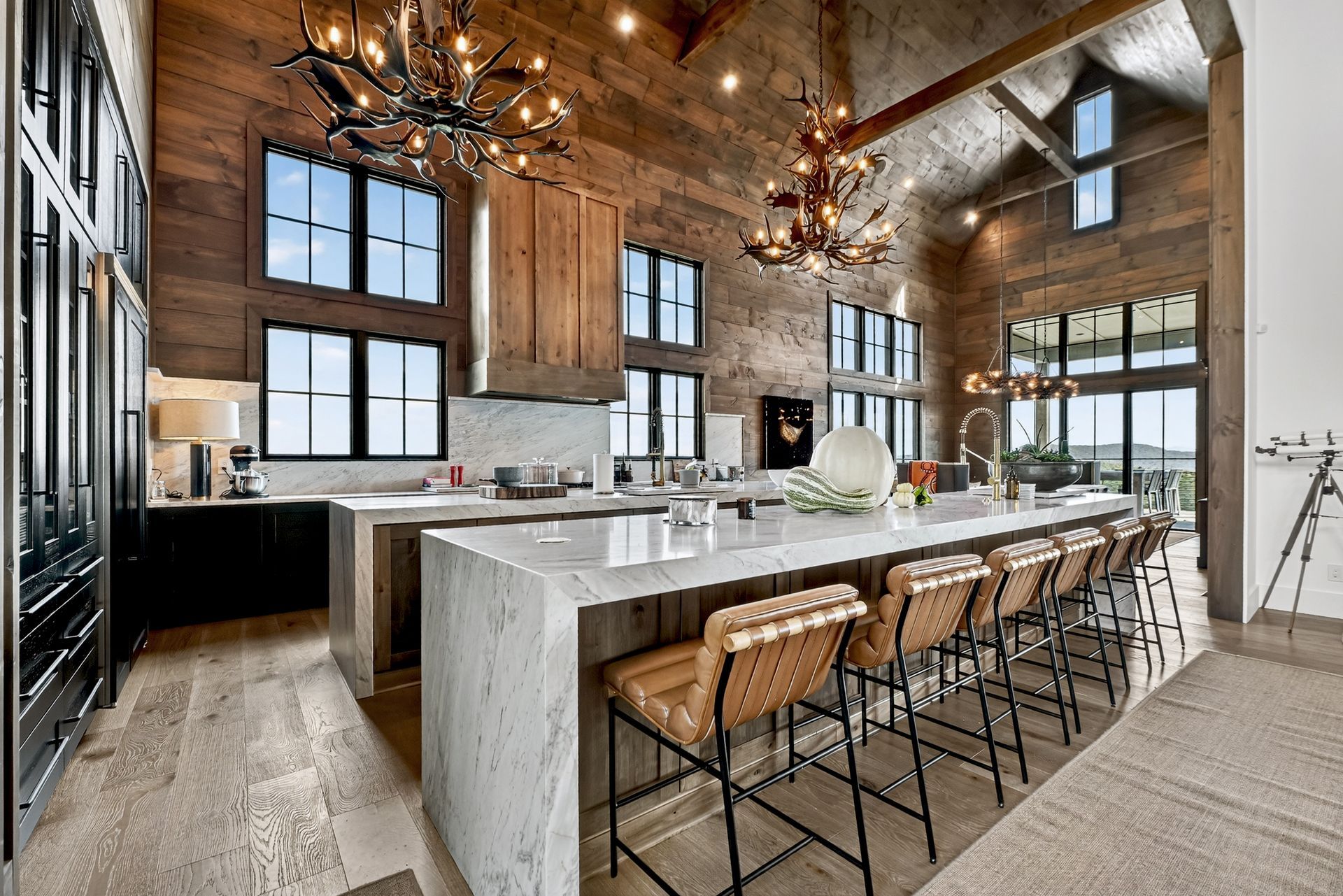 Kitchen with rustic wood walls, large island with seating, chandeliers, and windows overlooking a view.
