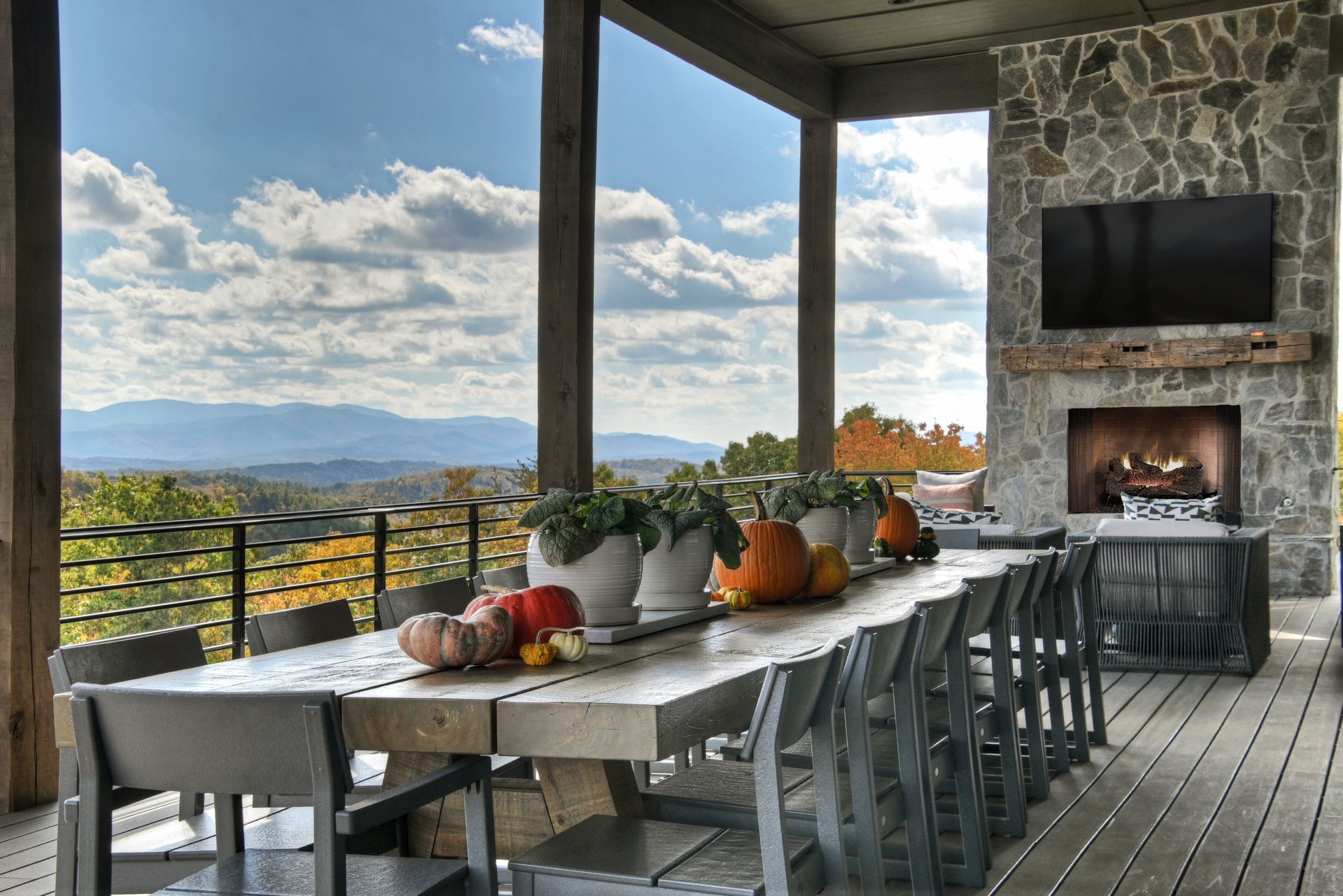 Outdoor dining area with long table, fireplace, TV, and mountain view, decorated with pumpkins.