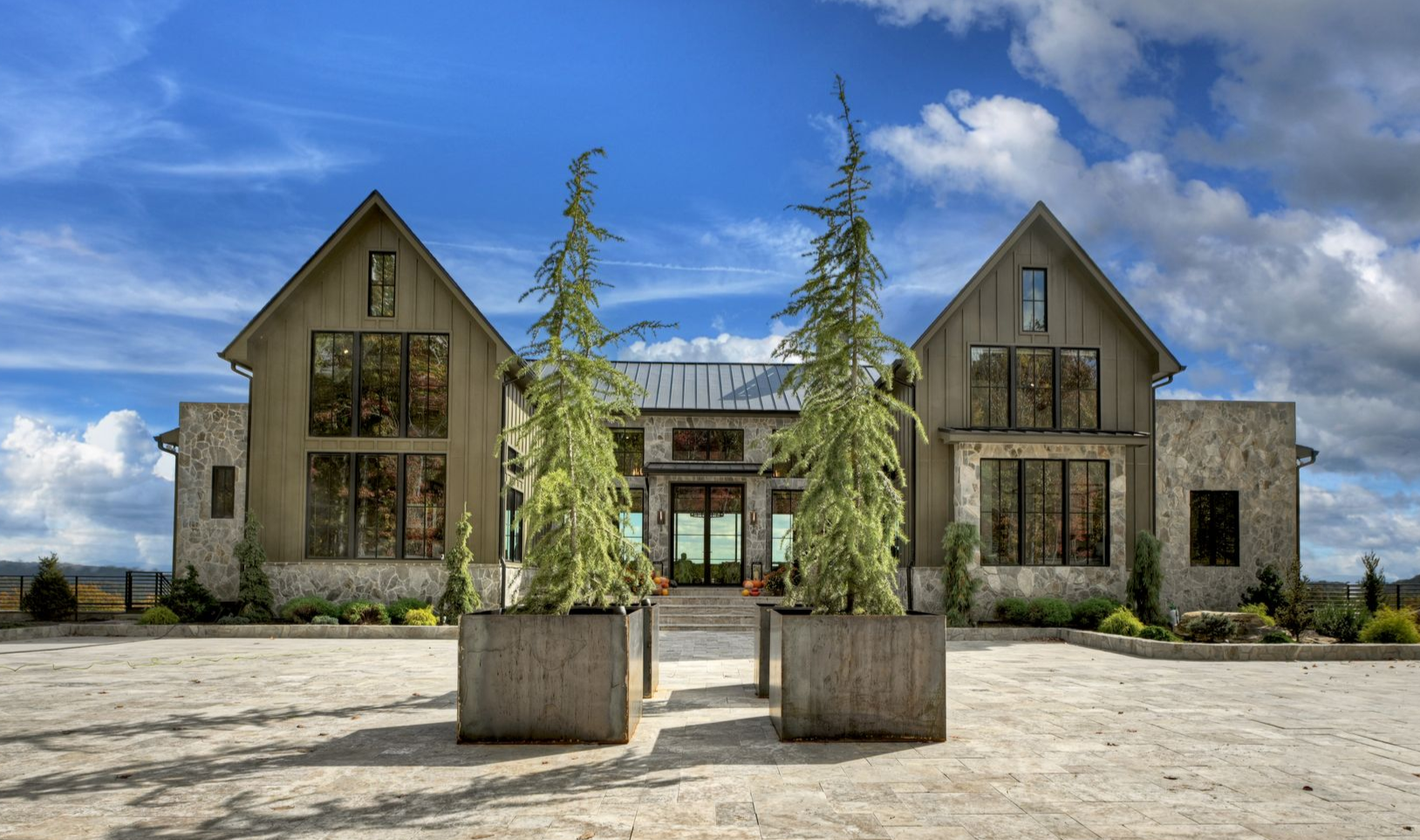 Modern farmhouse with stone accents, large windows, and two tall trees in square planters, against a blue sky.