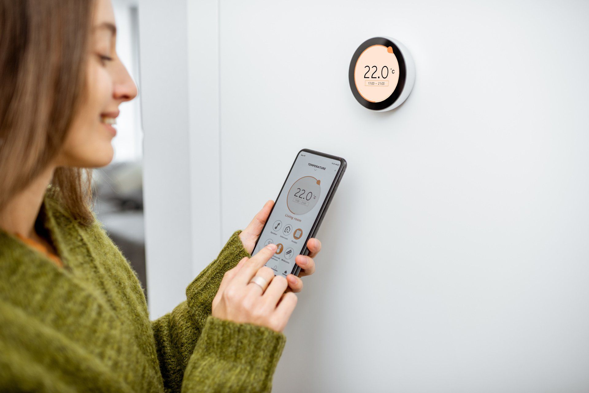 An individual adjusts the room temperature using a smartphone app, interacting with a wireless smart thermostat placed on a plain white background.
