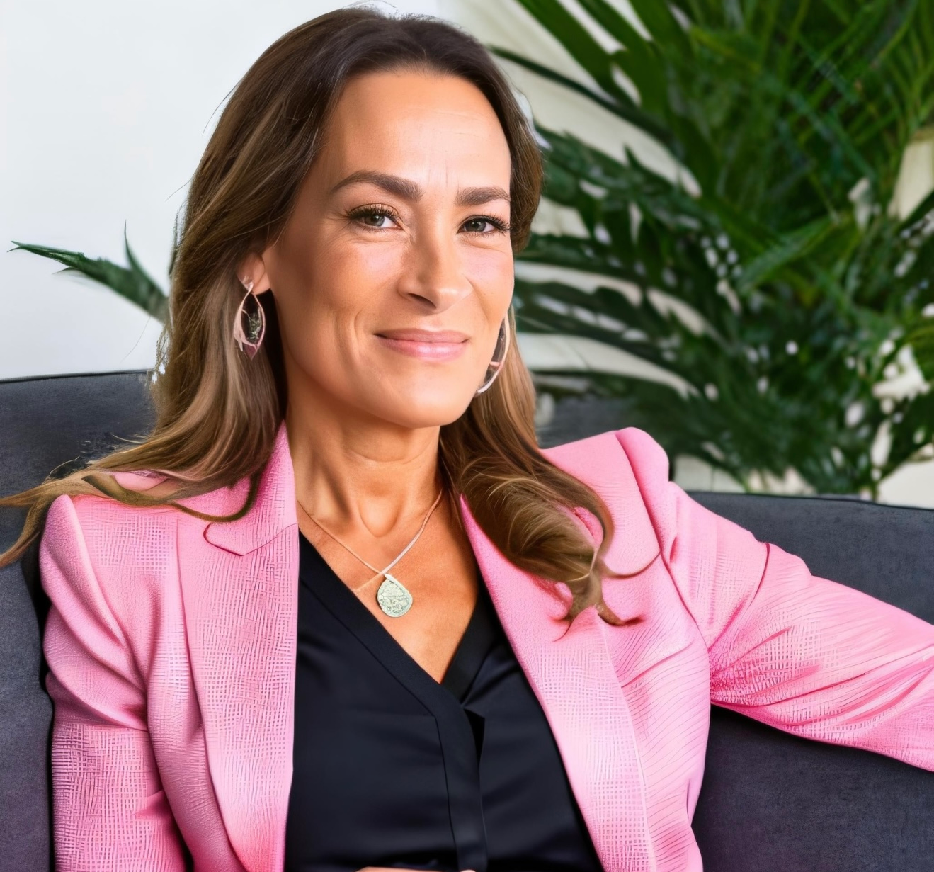Woman in pink blazer, smiling, seated on a gray couch, indoor setting.