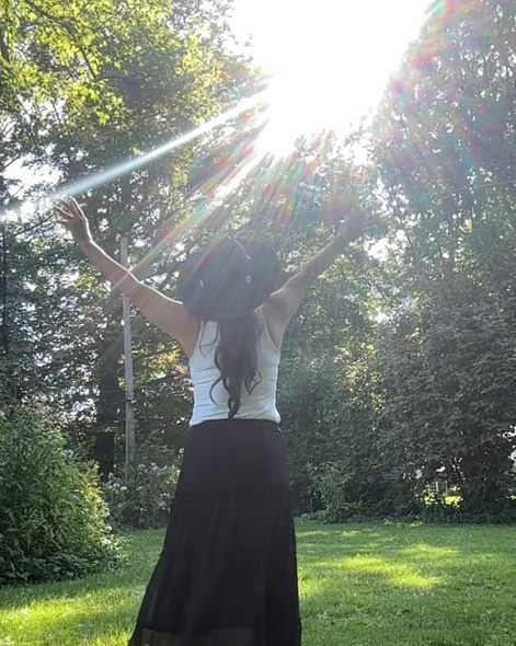 Woman with arms raised, standing in sunlight, wearing a white tank top, black skirt, and hat, in a green park.