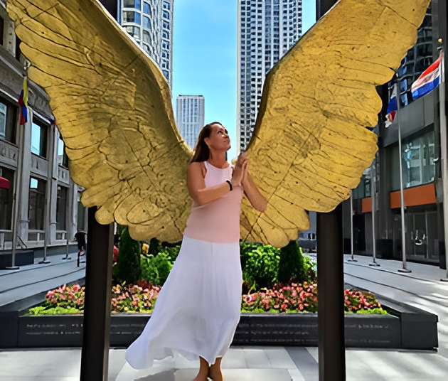 Woman poses with golden angel wings sculpture outdoors, hands clasped, wearing pink top and white skirt. City setting.