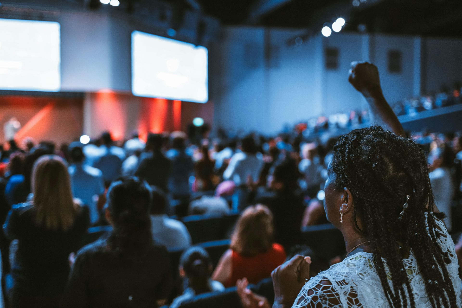 Woman with fist raised in a crowded auditorium with stage and screens lit up.