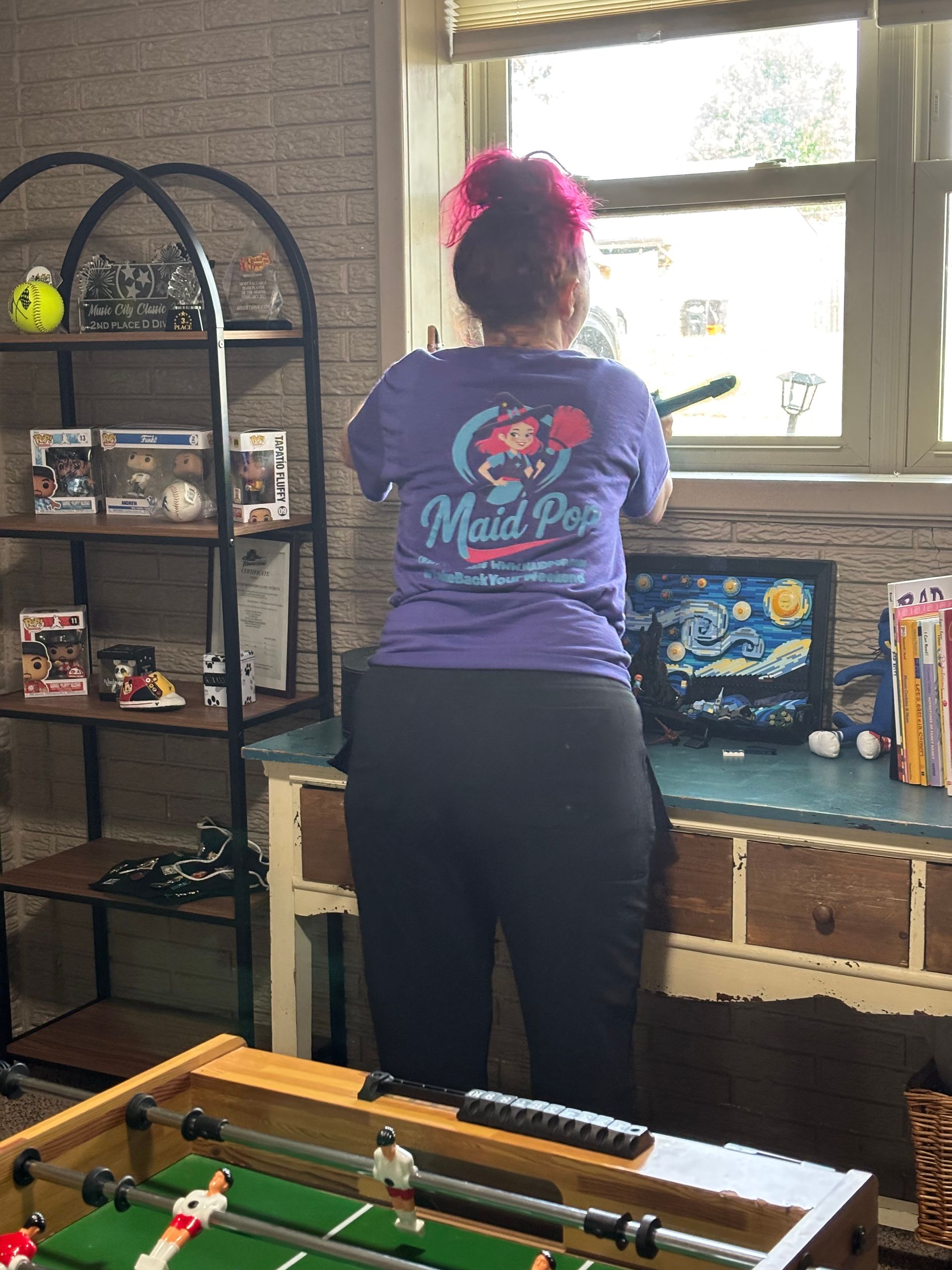 Woman in purple shirt, pink hair, looking out a window above a desk with a computer and bookshelf