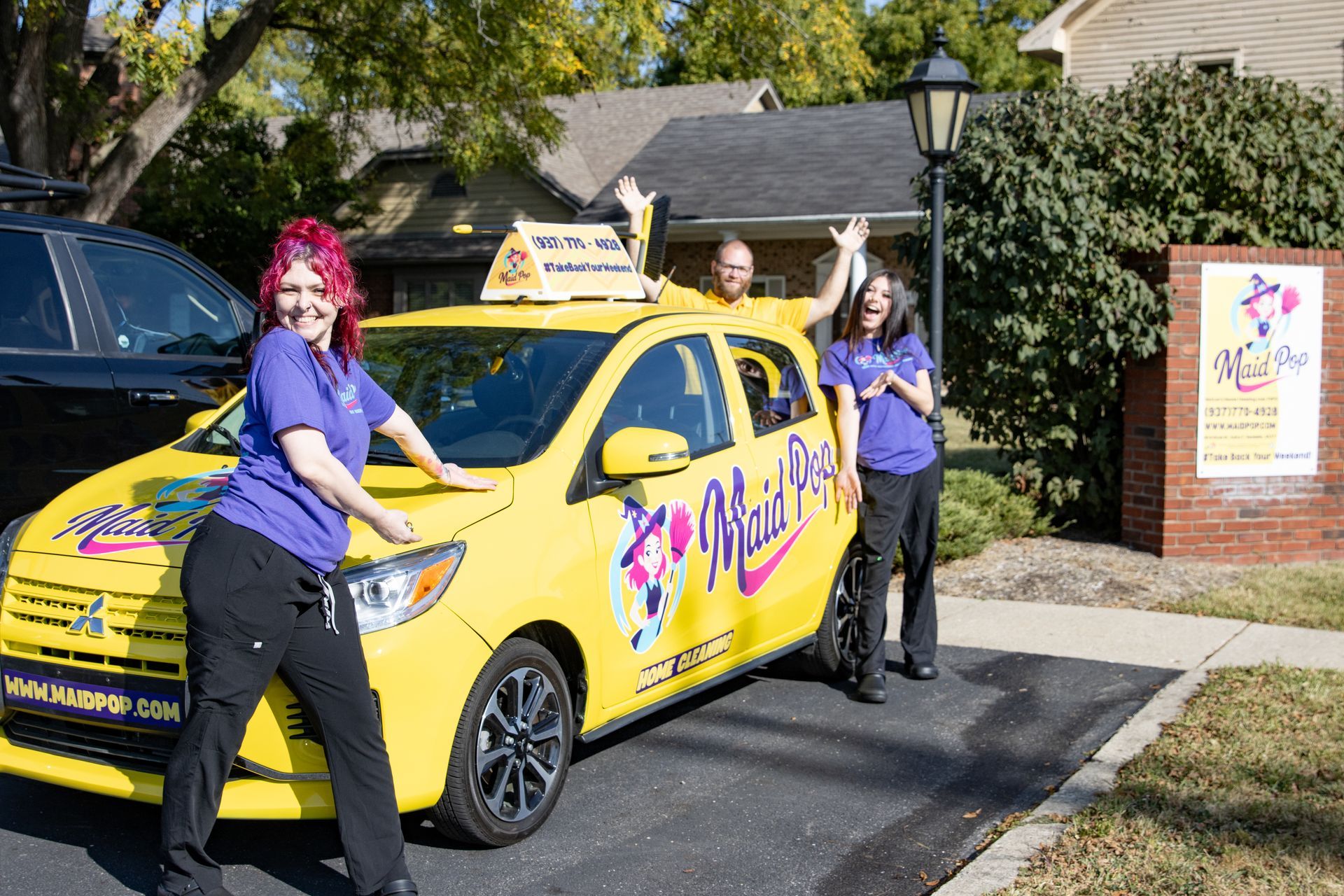 Three people pose with a yellow car, one person stands on the roof