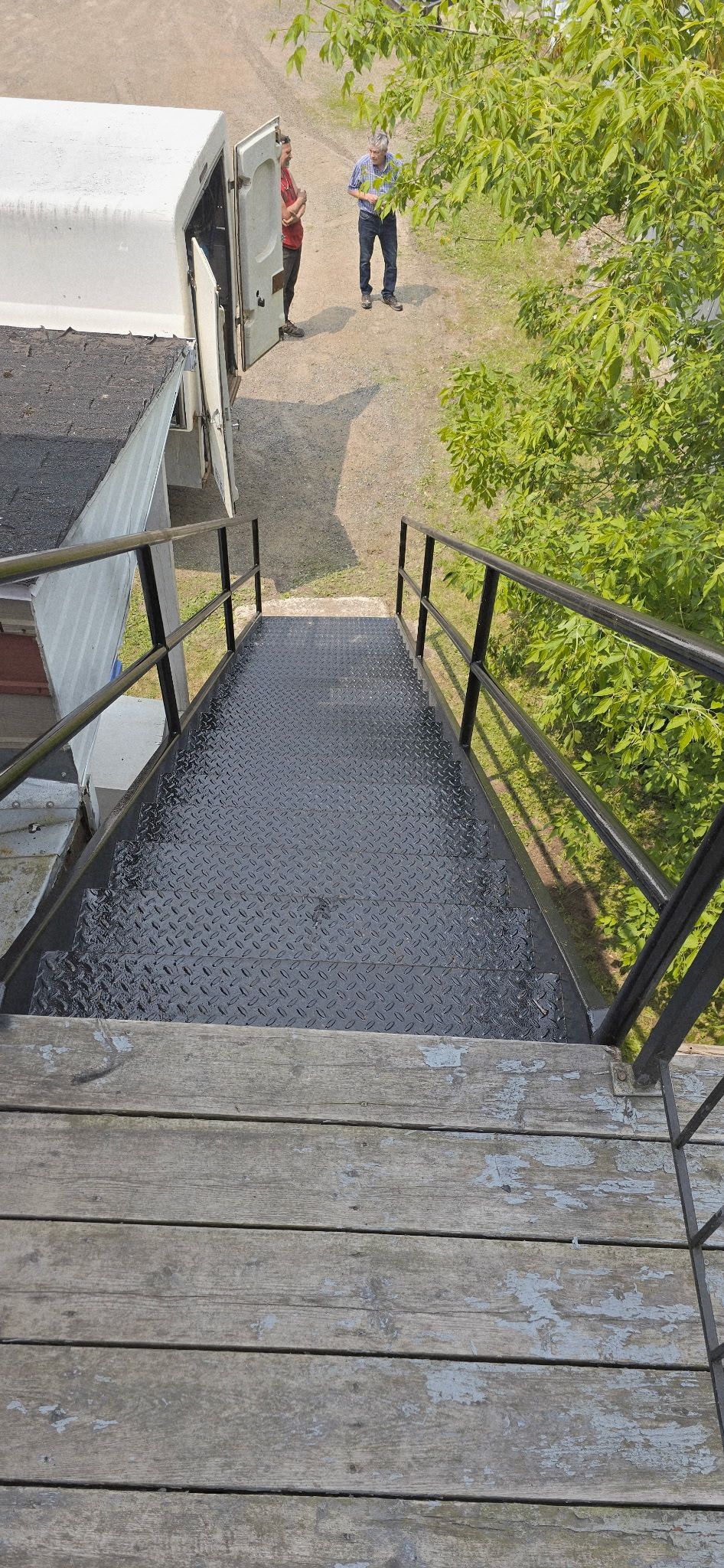Vue depuis un escalier en bois avec rampe en métal, vers un chemin de terre avec des personnages et un bâtiment.