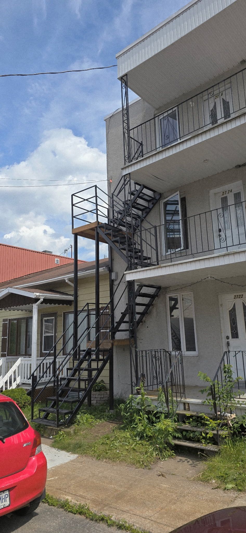 Un immeuble à plusieurs étages avec un escalier de secours à côté d'un autre bâtiment et d'une voiture rouge.