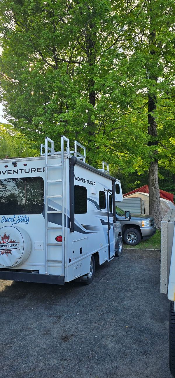Camping-car blanc stationné sur du gravier, entouré d'arbres et d'une camionnette de couleur claire.