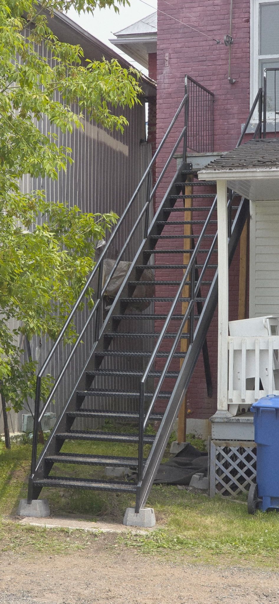 Un escalier de secours extérieur en métal contre un bâtiment en briques rouges et une maison blanche pâle.