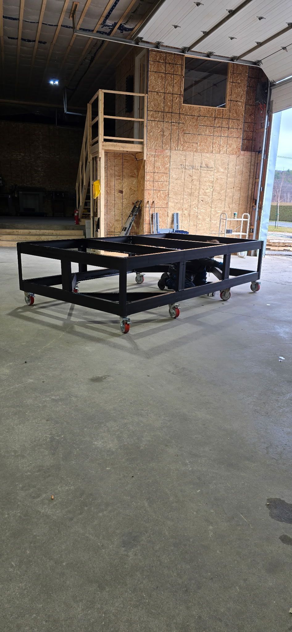 Plateforme roulante noire dans un atelier au sol en béton, avec une structure en briques et un échafaudage en arrière-plan.