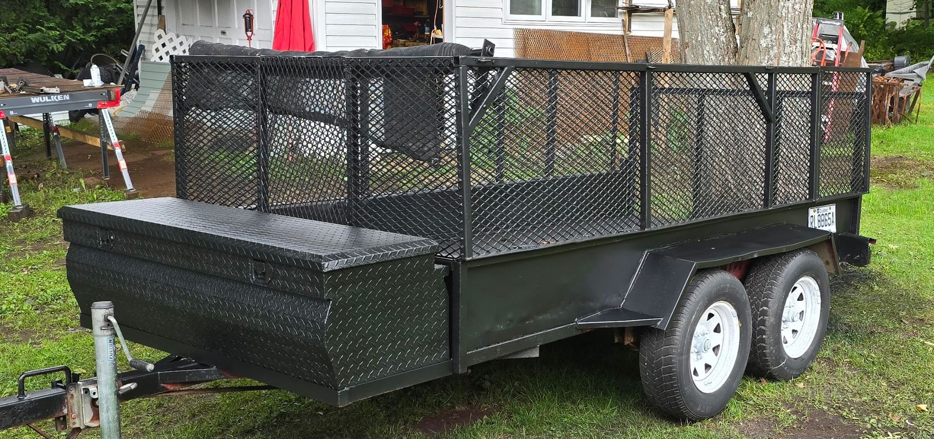 Une remorque utilitaire noire avec une enceinte grillagée et un coffre de rangement en tôle diamantée garée sur l'herbe.