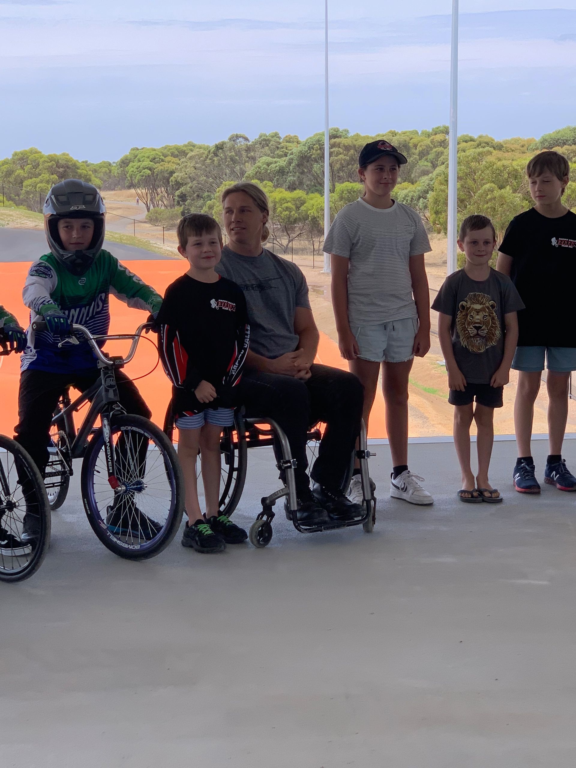 A man in a wheelchair is standing next to a group of children on bikes.