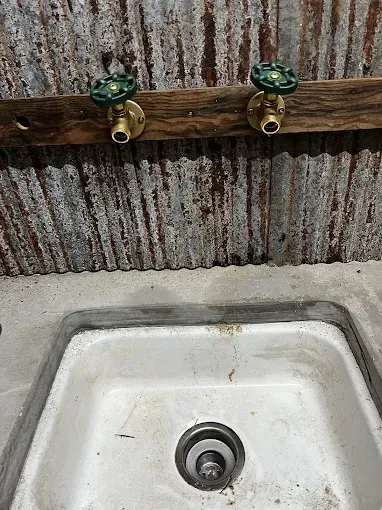 Bathroom sink with white basin, brass faucets with green handles, against corrugated metal wall.