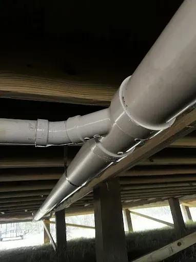White PVC pipes beneath a wooden structure, possibly a house, with a grass and post background.
