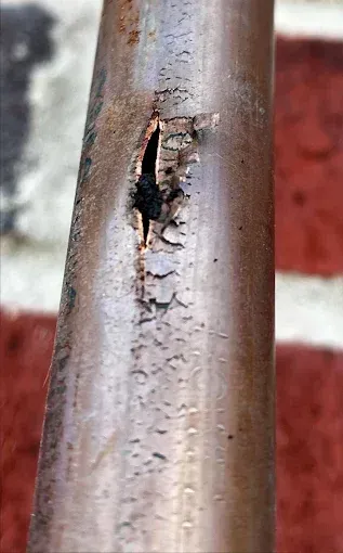 Damaged copper pipe with a split and flaky, brown material, against a blurred brick background.