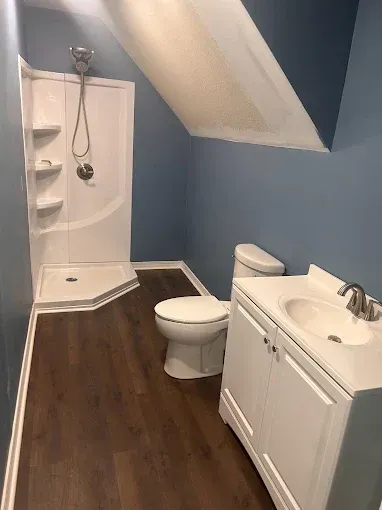 Small bathroom with white shower, toilet, and vanity. Blue walls, brown floor.