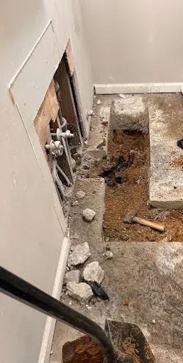 Hole in wall and floor showing plumbing; shovel, debris and concrete pieces in a room under construction.