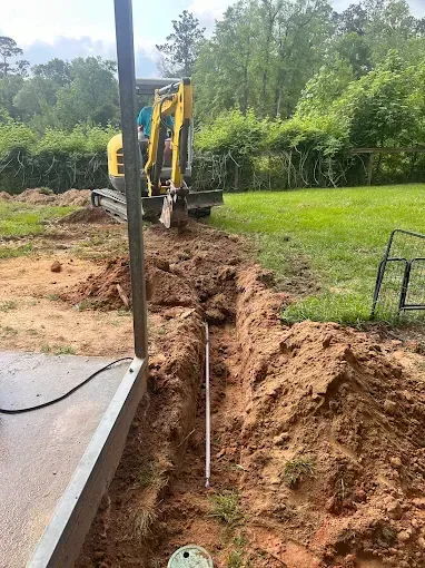 A small excavator digging a trench in a grassy yard, with a partially visible patio structure.