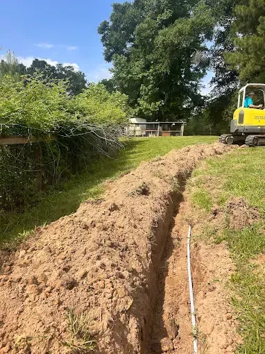 A long trench dug in a grassy yard; a small excavator and green foliage in the background.