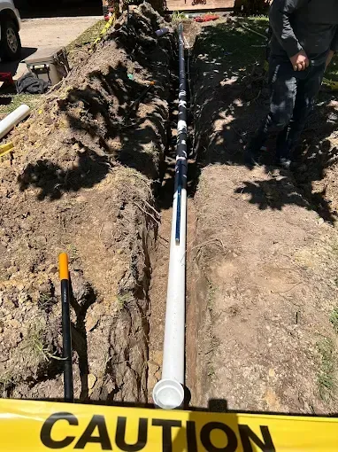 Trench with white pipe laid inside, caution tape, and a worker. Outdoors.