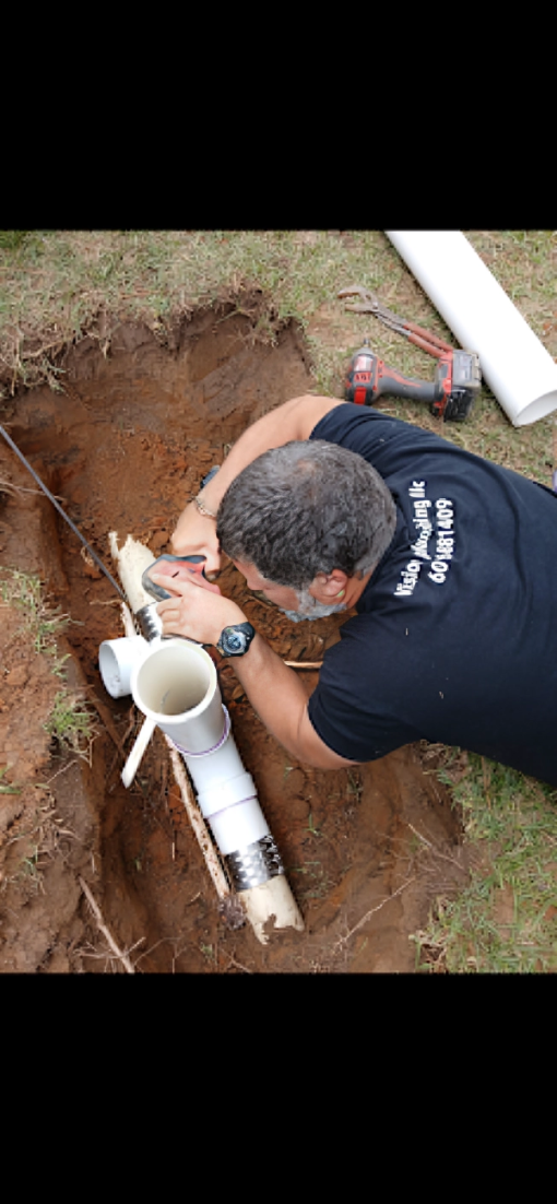 A man works on pipes in a dirt trench outdoors. He wears a black shirt and a watch.