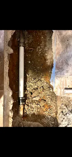 A white pipe exposed in a trench dug into a floor. The trench is filled with soil and rock.
