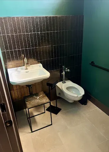 A bathroom with a white sink and toilet, gray floor, and black tiled wall. A small stool sits near the sink.