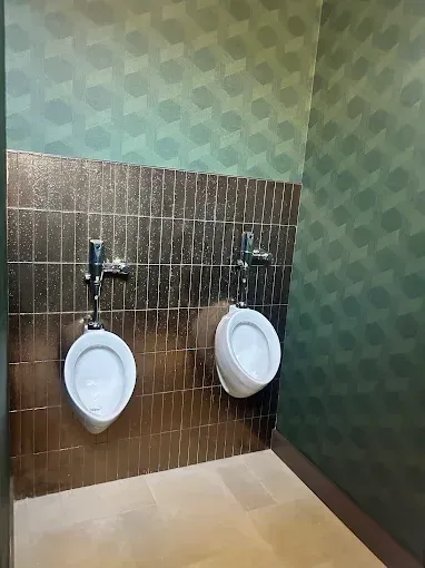 Two urinals on a gold tile wall, against green patterned wallpaper.