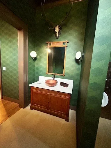 Bathroom with green patterned wallpaper, a wood vanity, and a deer skull above the mirror.