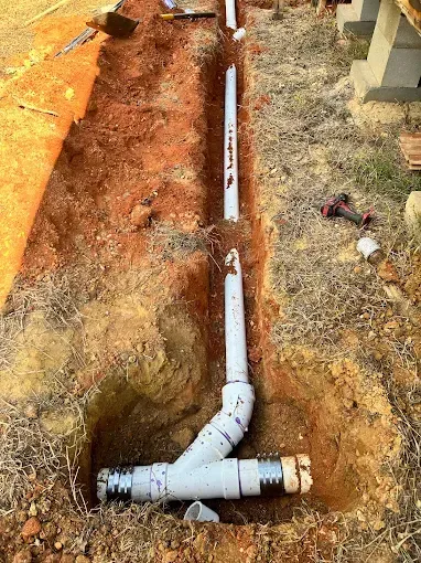 Trench with white plumbing pipes being installed in the ground. A drill and tools are visible.
