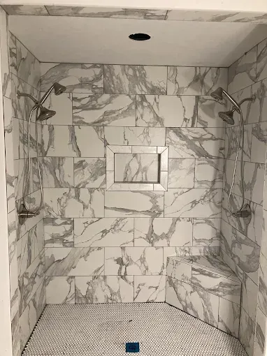 Marble-tiled shower with two shower heads, recessed shelf, and small seating area; white and gray.