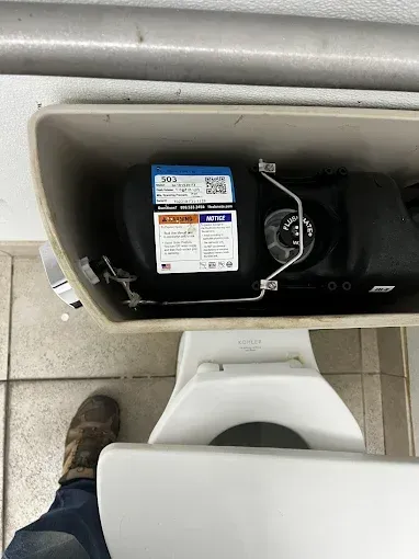 Toilet tank interior showing a black fill valve with blue label, metal hardware, and part of a leg and shoe.