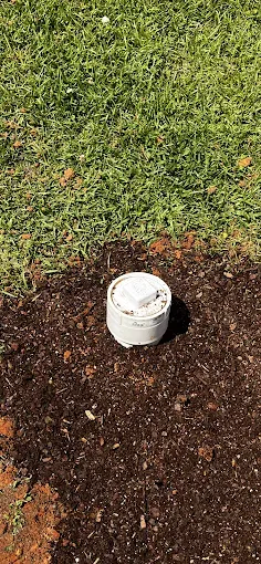 A white capped cylinder is set in brown soil next to green grass.