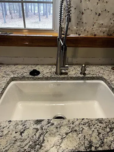 Kitchen sink with granite countertop, stainless steel faucet, soap dispenser, and window in the background.