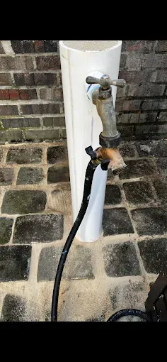 An outdoor water faucet with a black hose attached, mounted on a white pipe against a brick wall.