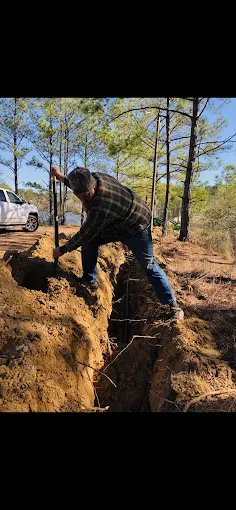 Man digging a trench outdoors near a vehicle and trees on a sunny day.
