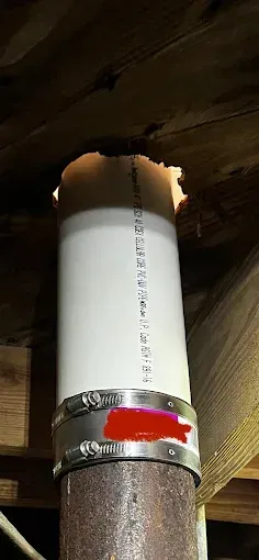 White PVC pipe connected to a rusty pipe with a metal clamp, passing through wooden ceiling.