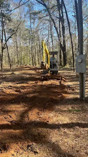 An excavator digs a trench in a wooded area next to a utility pole.